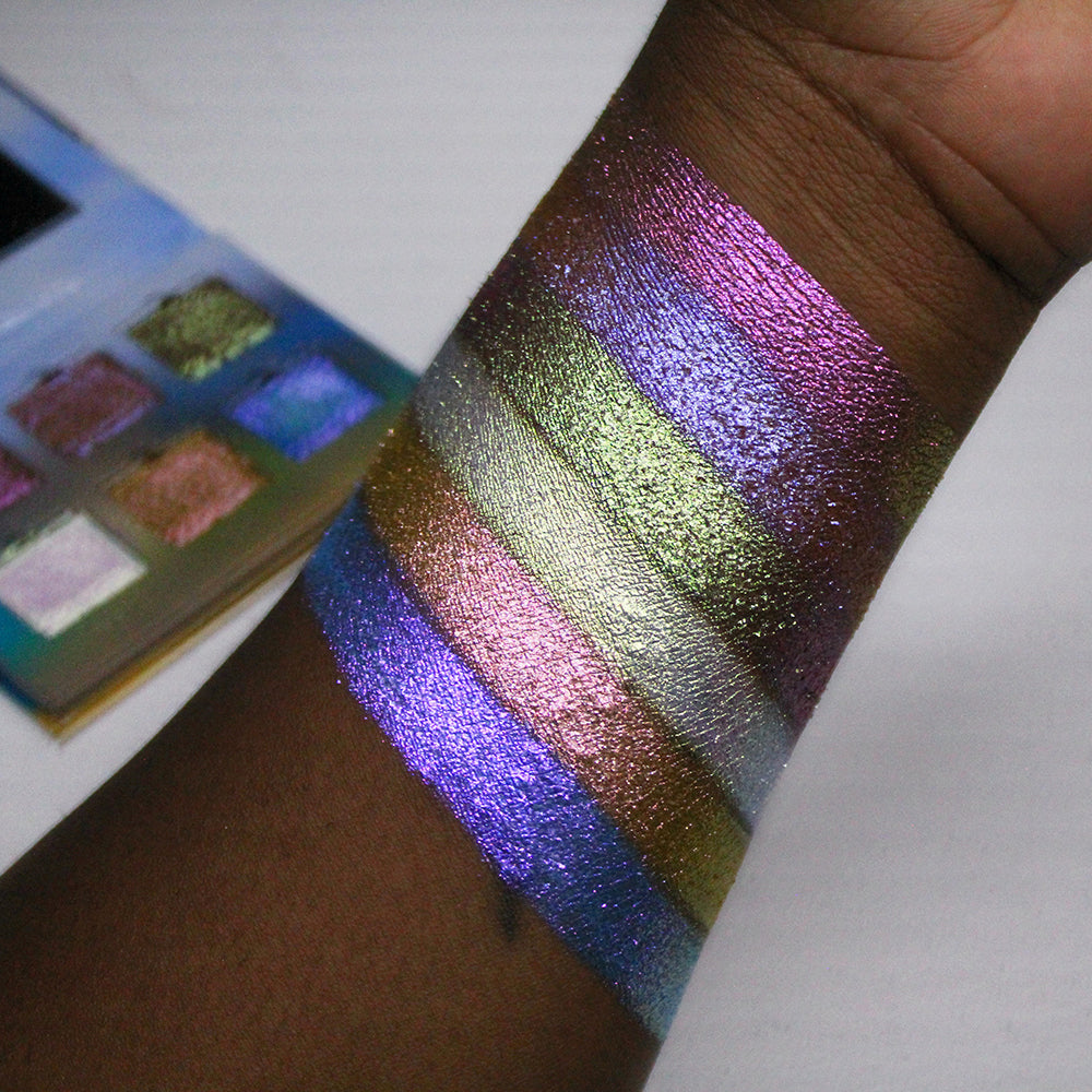 Arm swatches of all six Galápagos Palette shadows on deep skin tone. Palette propped open in the background.
