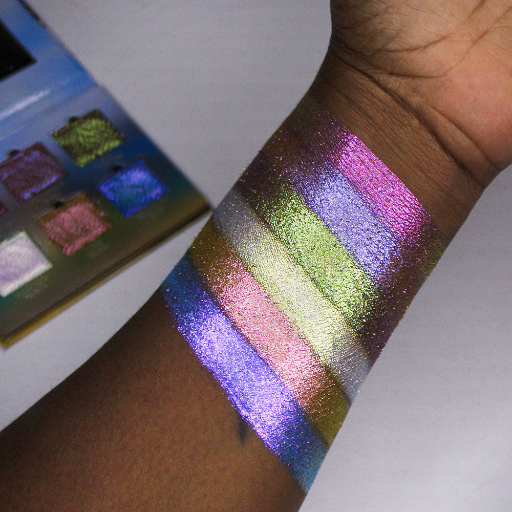 Arm swatches of all six Galápagos Palette shadows on deep skin tone, with palette propped open in background.